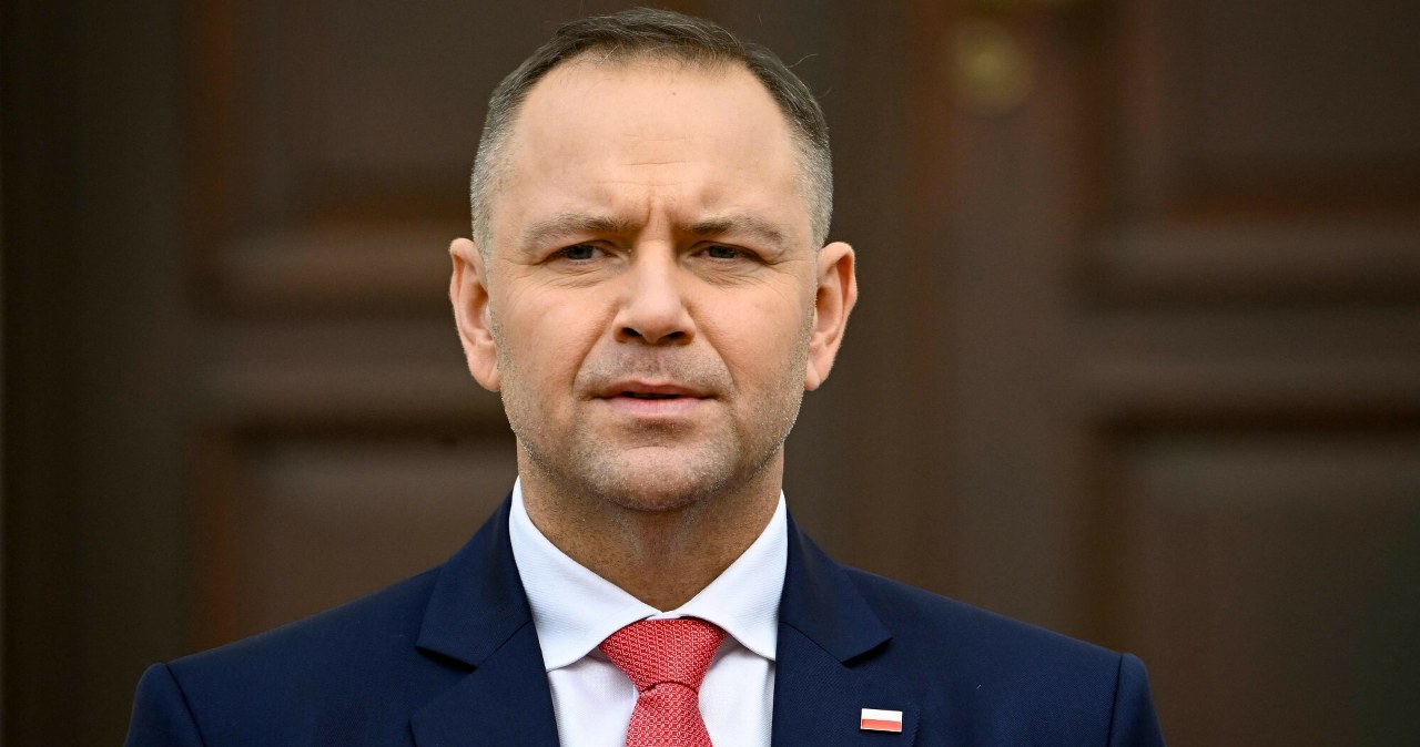 Prezydent Karol Nawrocki w ciemnogranatowym garniturze i czerwonym krawacie na tle brązowego tła