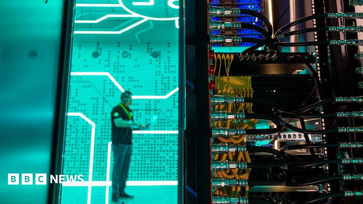 A man standing against a bright green background in a data centre - in the foreground is the back of a large computer with