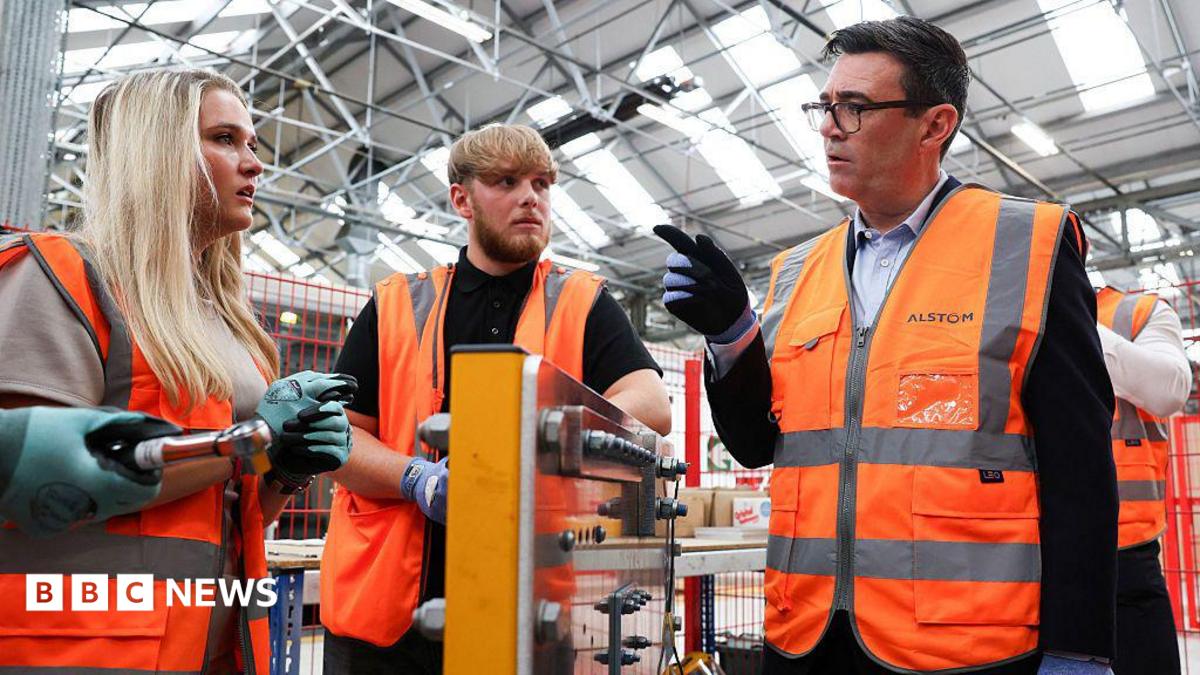 Andy Burhnam wears a hi-viz jacket while talking to two young workers at a factory, also wearing hi-viz jackets, one female