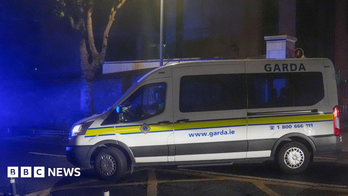 A white Garda van with a yellow stripe along its side pulls out onto a main road on a dark night