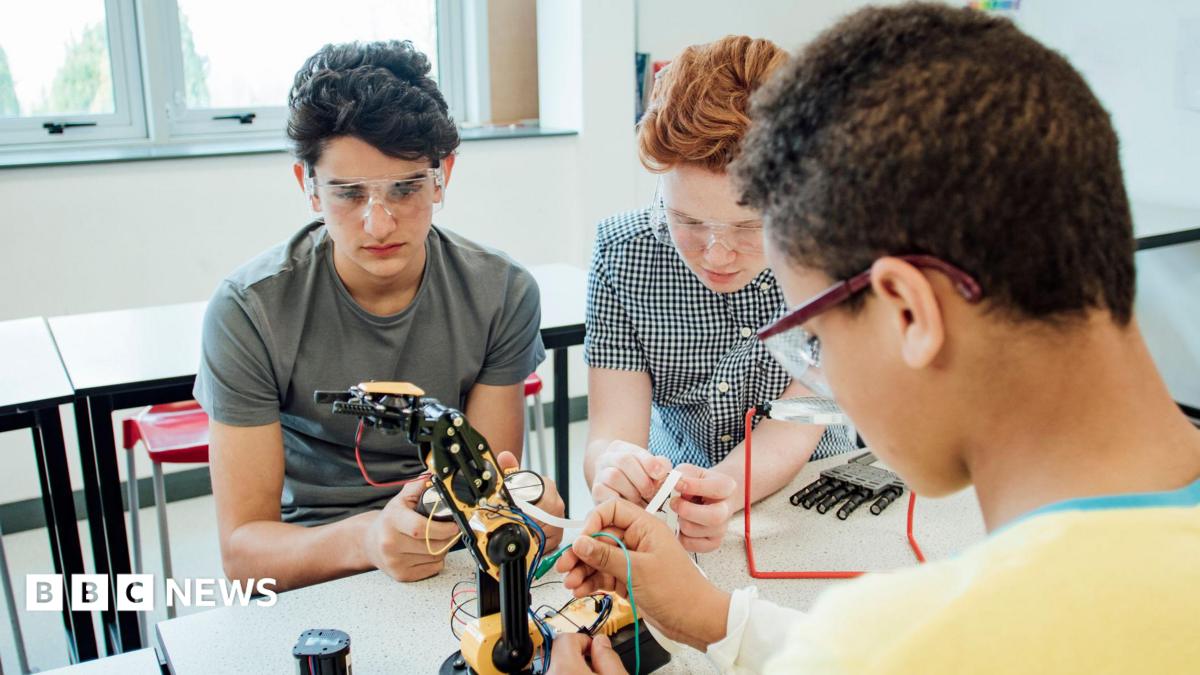 Three students wearing safety goggles work together on a robotic arm in a classroom or laboratory, connecting wires and