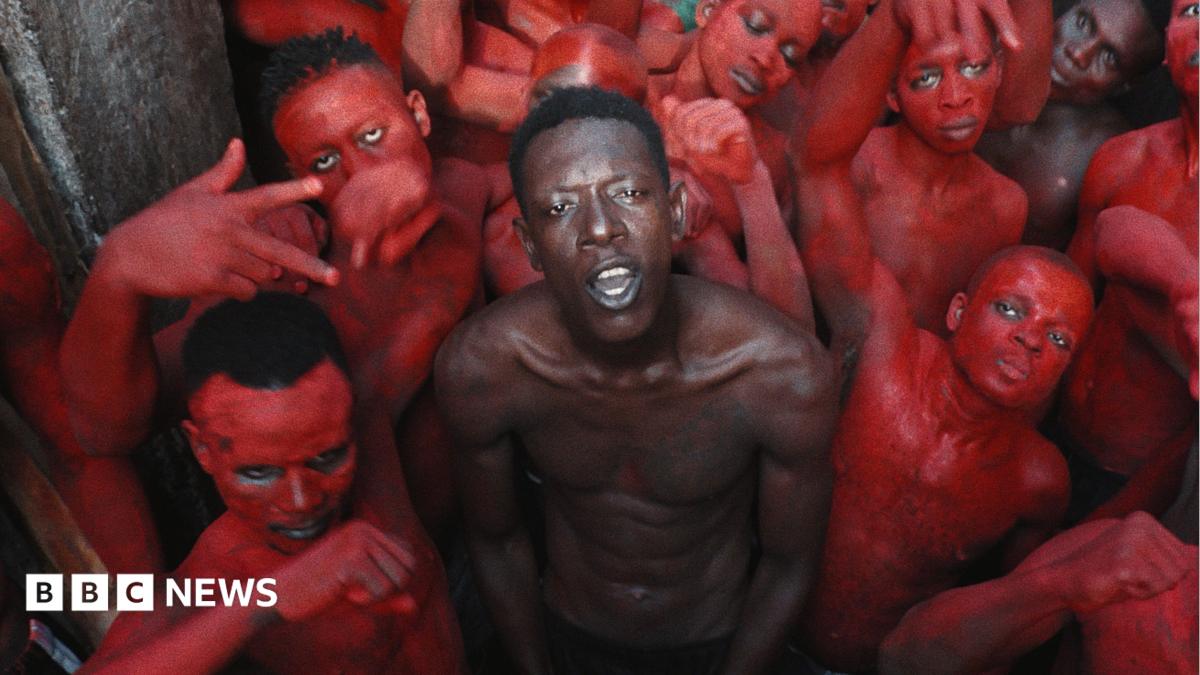 A topless Empereur stands in the centre of a group of young men, who are painted in red
