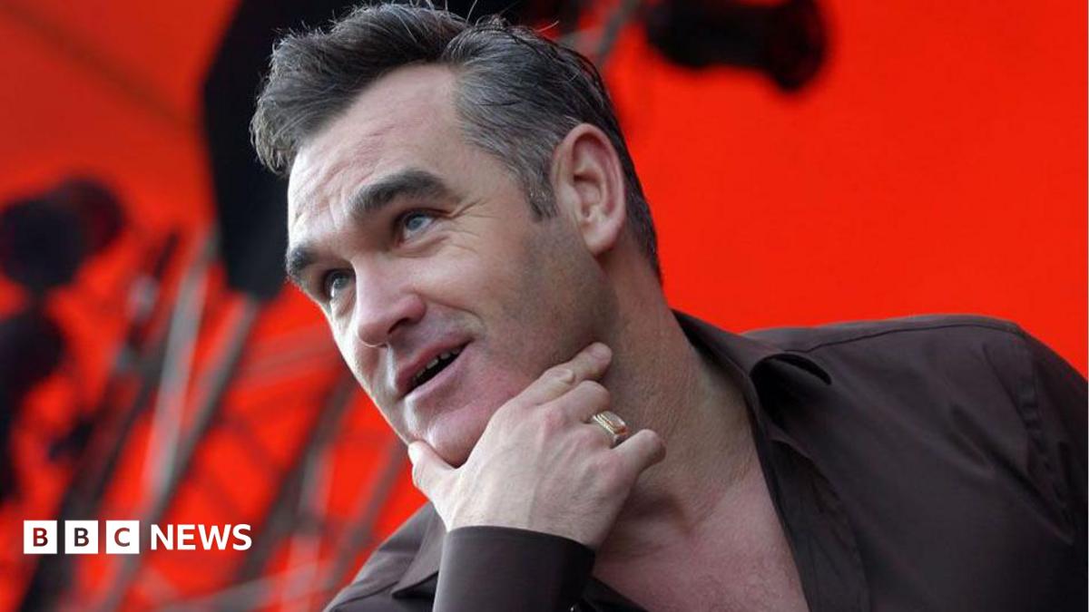 Head and shoulders of Morrissey on stage with his right hand thumb and index finger cupping his chin. He is wearing a gold