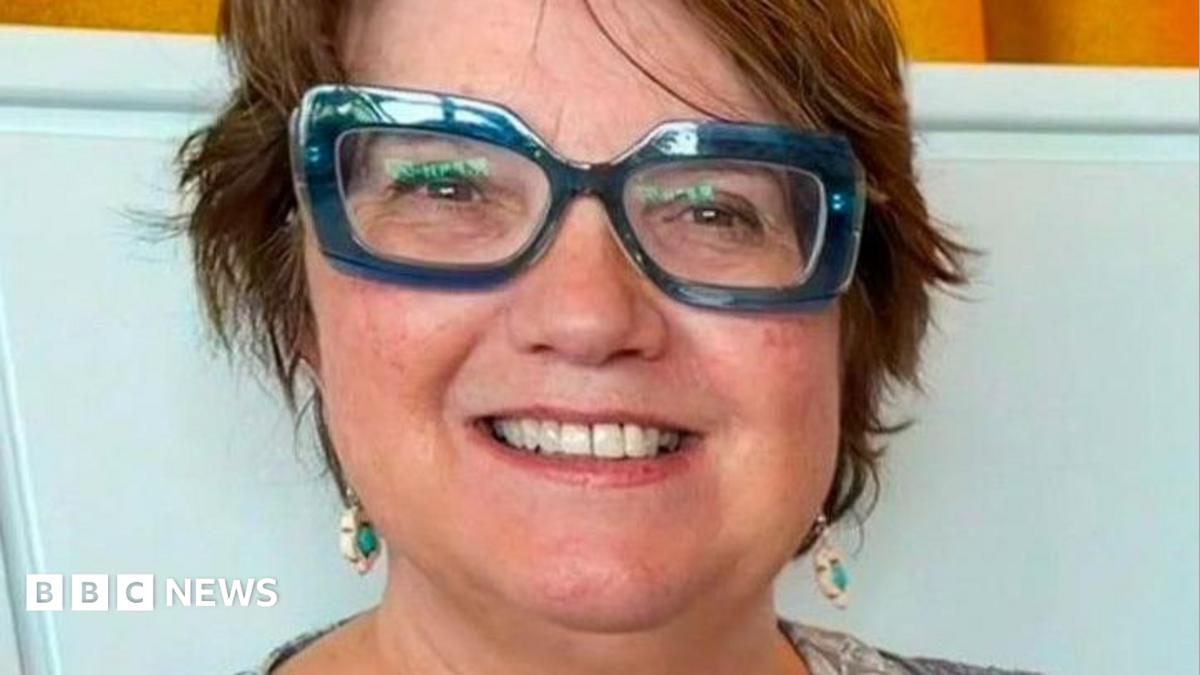 The photo shows a person with short brown hair wearing large blue-framed glasses. They are dressed in a light blue patterned