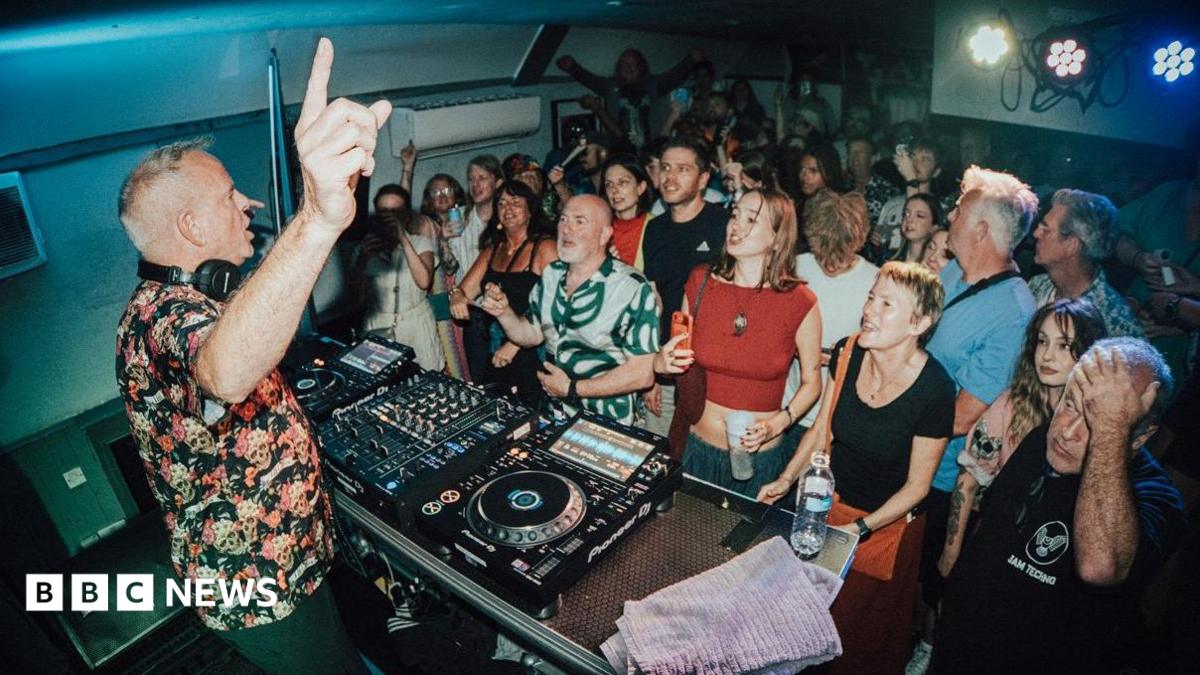 DJ Fatboy Slim points upward while performing behind a mixing deck in a compact room filled with a tightly packed audience
