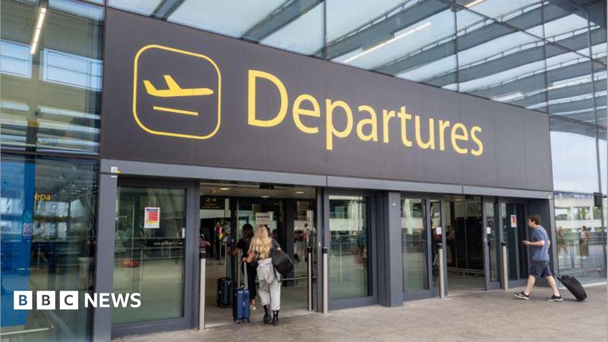 The departures entrance at Gatwick Airport with large yellow "Departures" signage above glass doors. Travellers with luggage