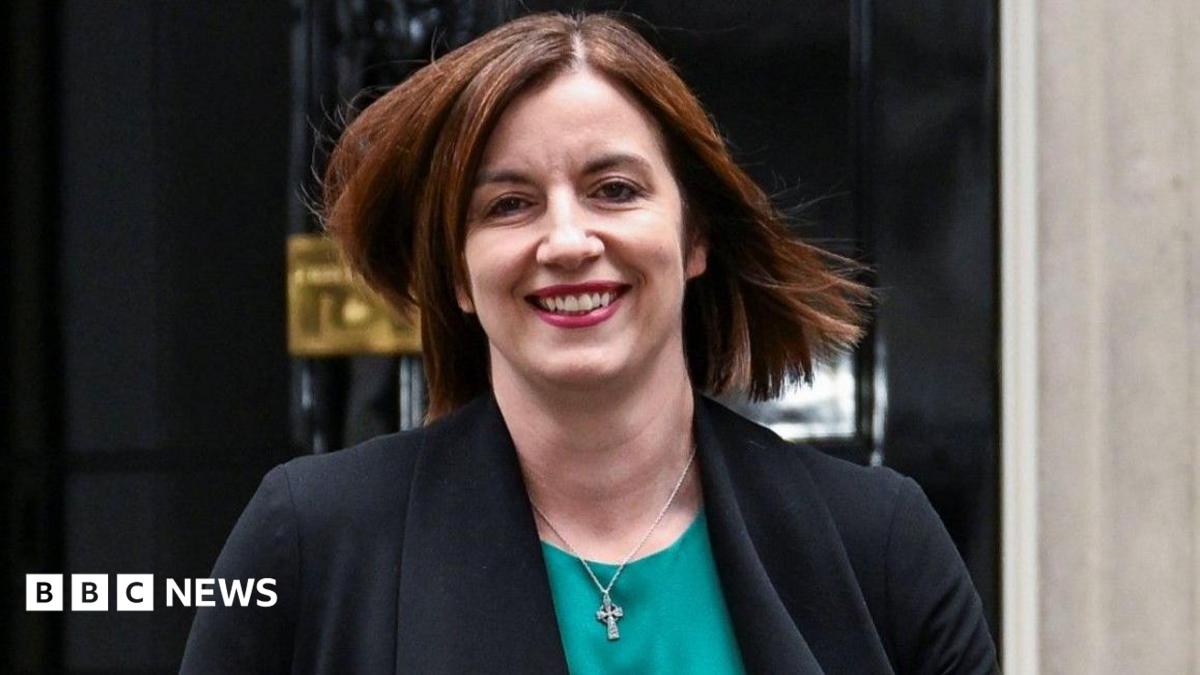 Phillipson wears a teal scoop-necked top with a black jacket and a small silver Celtic cross. She wears dark pink lipstick