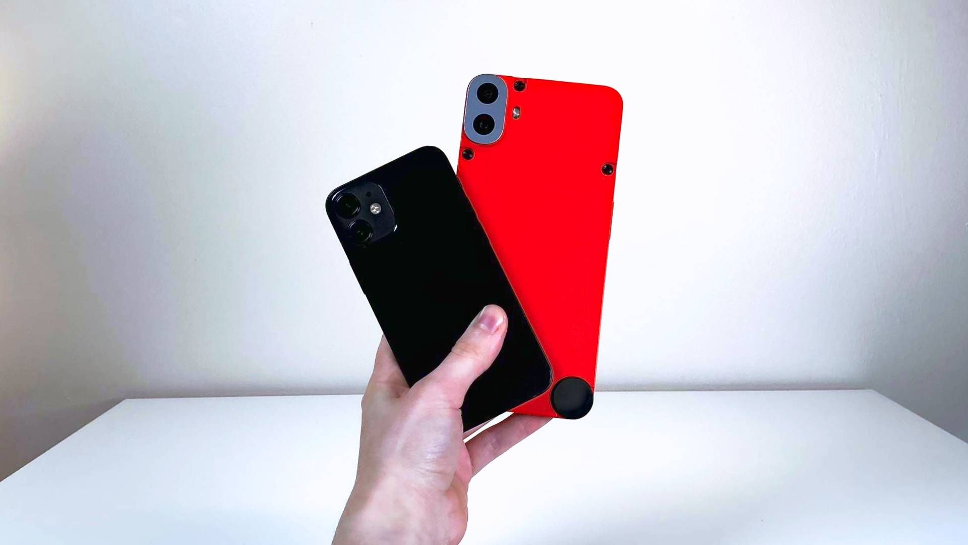 A hand holding the iPhone 12 Mini and CMF Phone 1 in front of a white table