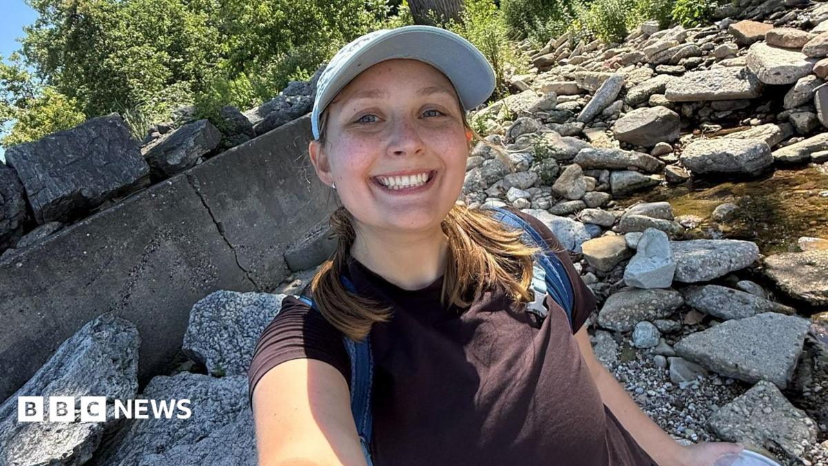 Megan Szostak stands in a rocky stream, taking a selfie and smiling at the camera. She wears a black t-shirt, blue backpack