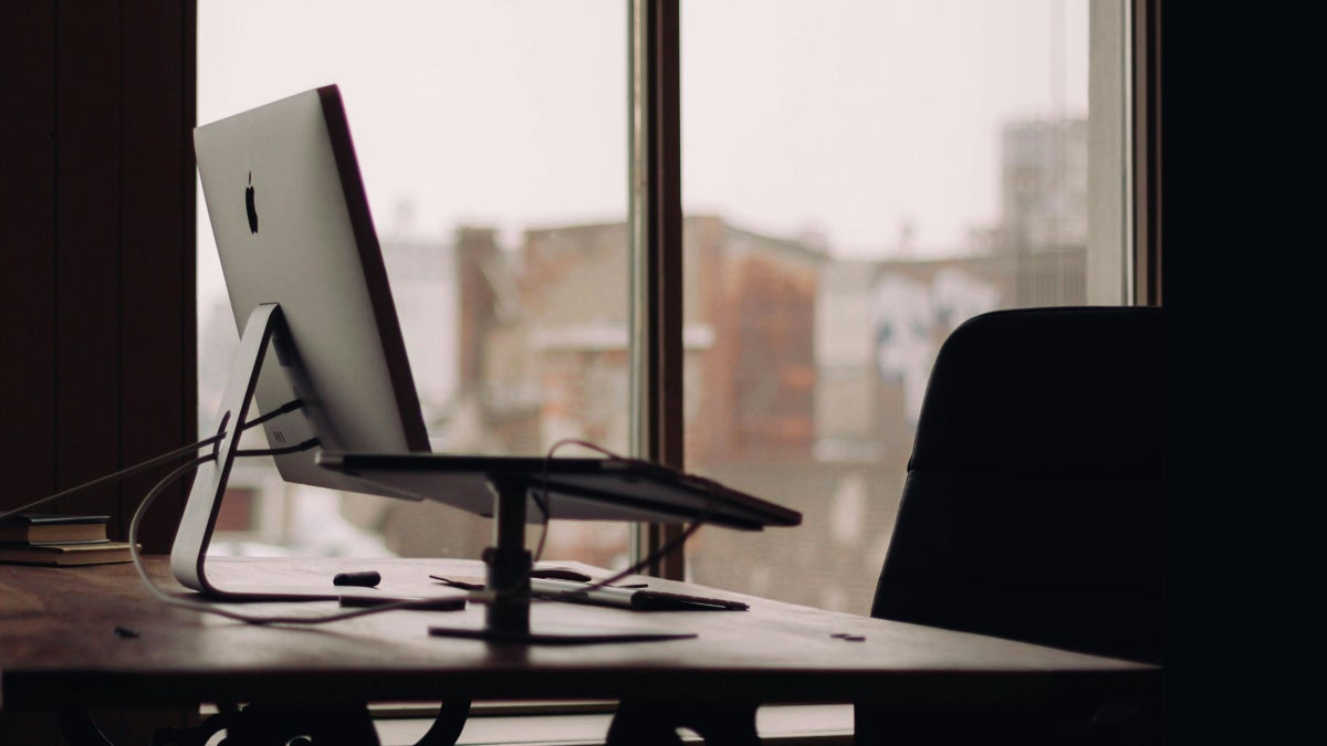 Close-up of an Apple iMac computer and desk with books and a laptop stand, with a dark chair in the foreground and a blurred