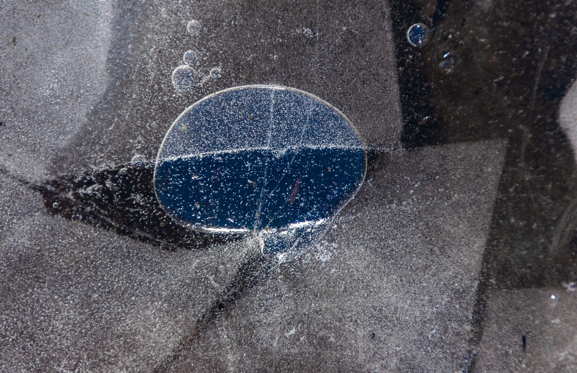 A circular pocket of dark blue water visible beneath a frozen, bubble-filled surface