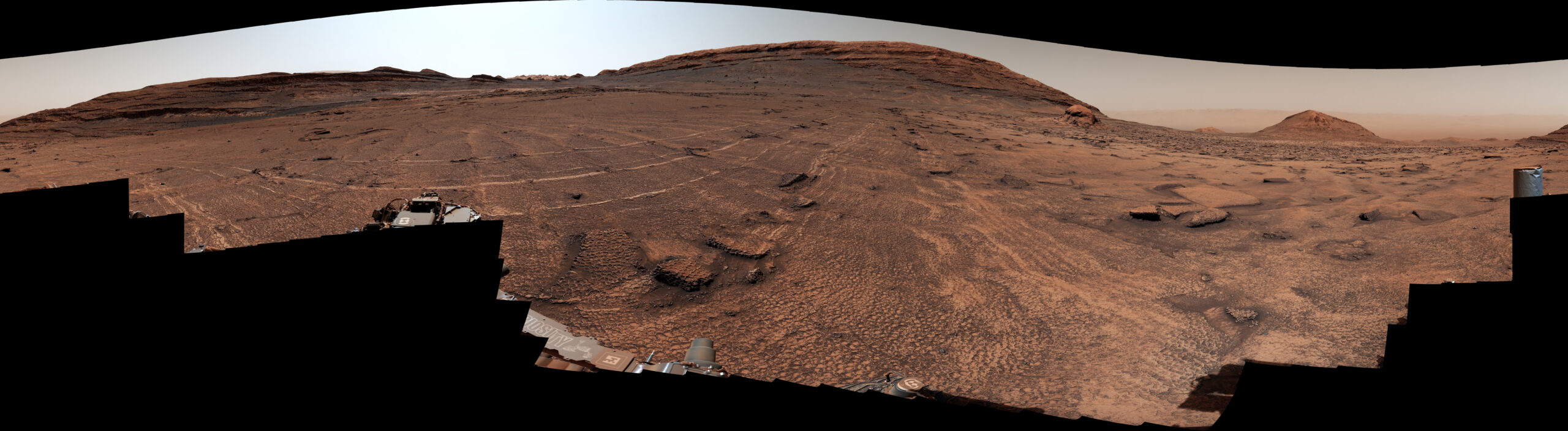 A panoramic view of a sweeping Martian plain covered in patterned fractures leading toward distant hills showcases the