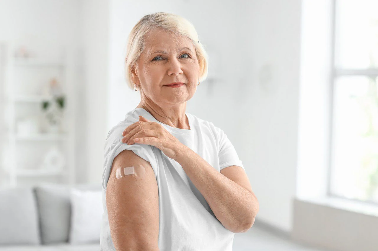 Older woman in a white shirt shows a small bandage on her upper arm after receiving a vaccination
