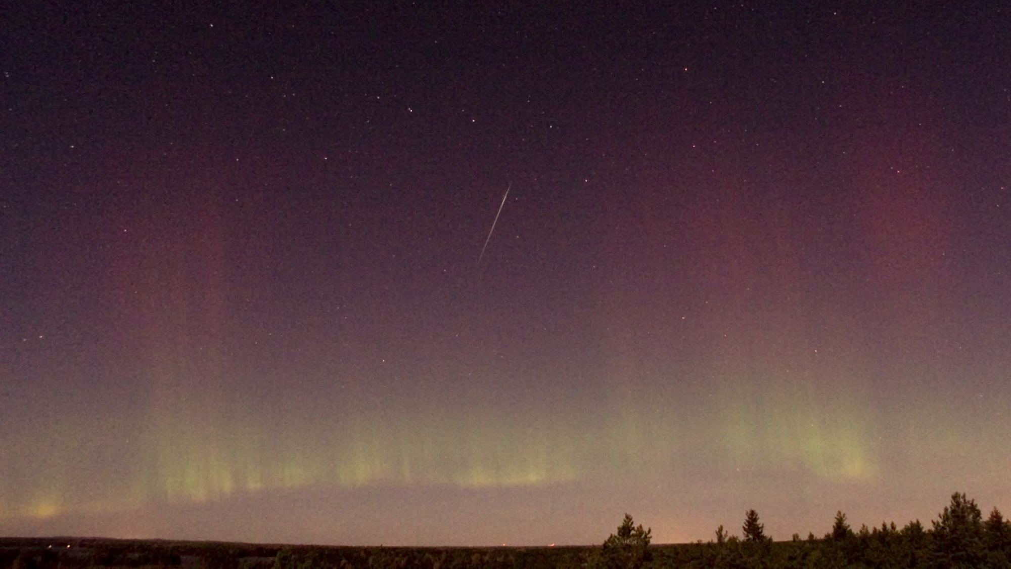 A lone shooting star streaks through a night sky riven with colorful aurora